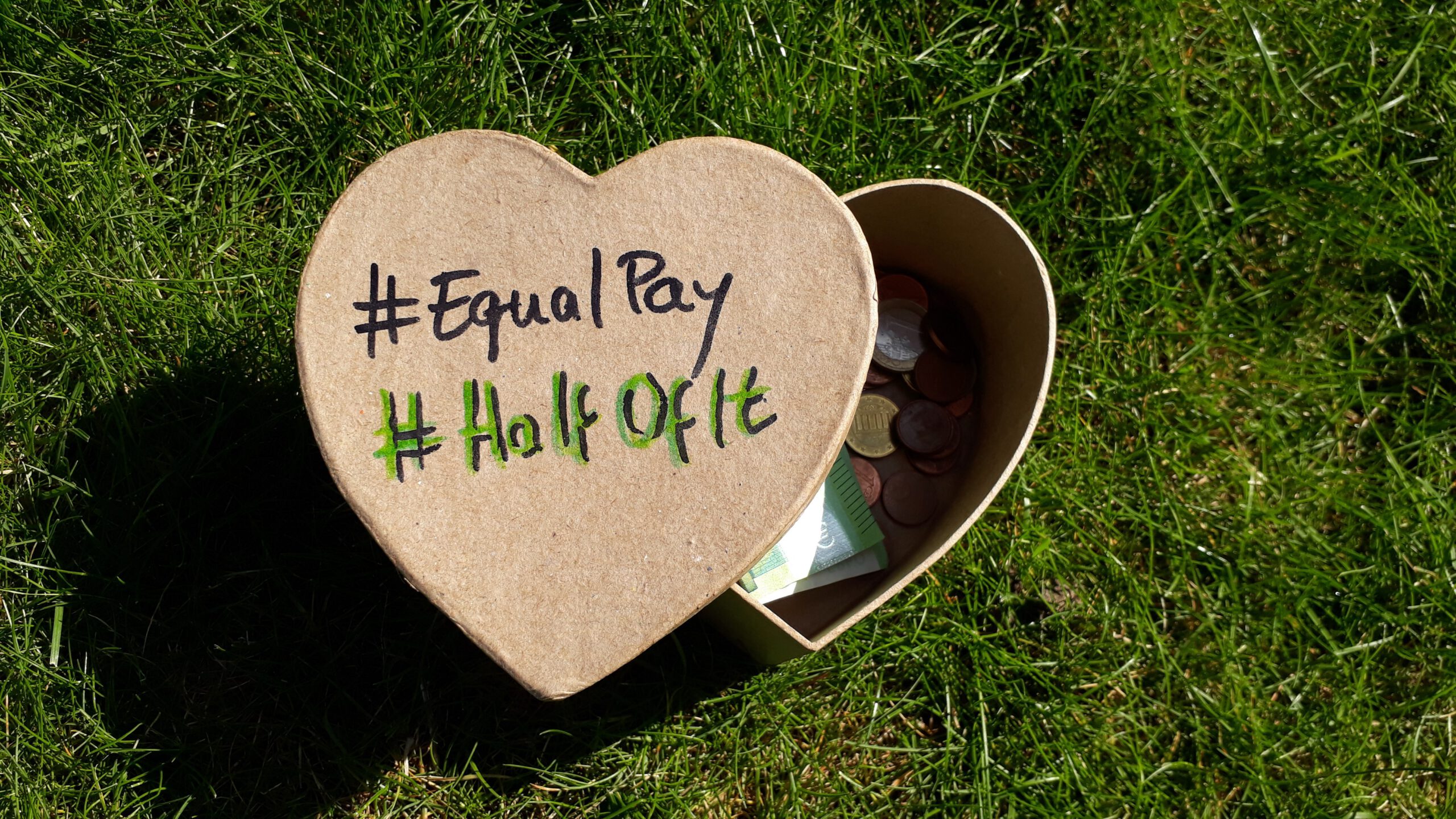 Foto mit einer Herzförmigen Kiste, die im Gras liegt und auf der #Halfofit und #EqualPay steht